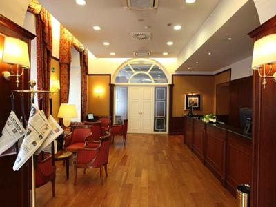 Jasna i przytulna lobby hotelowa z drewnianą podłogą, czerwonymi fotelami i ciepłymi lampami ściennymi.