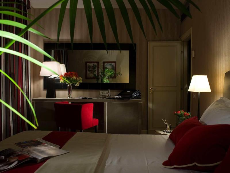 Pohodlný hotelový pokoj s postelí, psacím stolem, červenou židlí a tlumeným osvětlením.