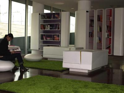 Moderne Hotellobby mit Bücherregalen, weißen Sitzmöbeln und grünen Teppichen, Person liest Buch.