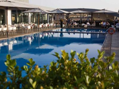 Großer blauer Außenpool mit umliegenden weißen Stühlen und Tischen vor einem modernen Gebäude.