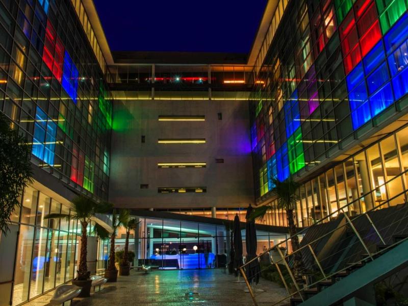 Modernes Gebäude mit Glasfronten, beleuchtet in Rot, Grün, Blau, bei Nacht unter dunklem Himmel.