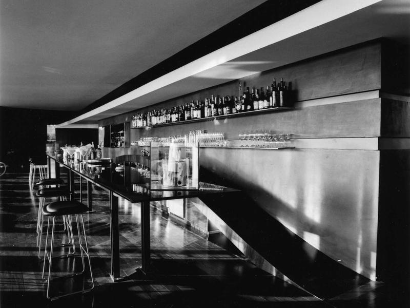 Schwarz-weiß Bar mit langen Tresen, Regal mit Flaschen und Hockern in modernen Linien und klaren Formen.