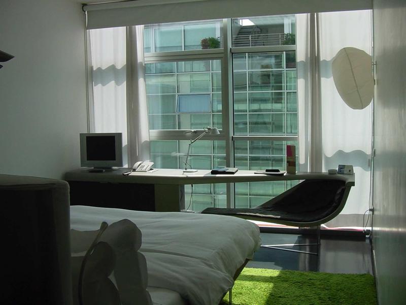 Modernes Schlafzimmer mit großem Fenster, Schreibtisch, Computer und schwarzem Sessel.