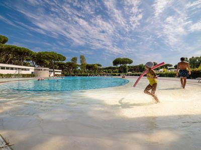 Deux enfants jouent au bord d'une grande piscine extérieure sous un ciel bleu avec des nuages.