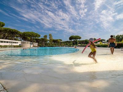 Des enfants jouent dans la partie peu profonde d'une grande piscine extérieure sous un ciel bleu avec des nuages.
