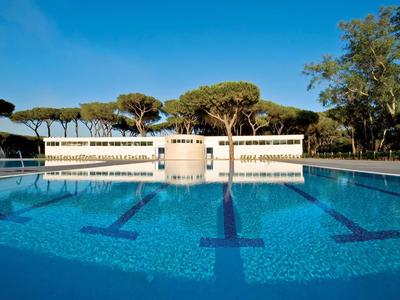 Une piscine extérieure avec de l'eau claire devant un mur blanc et des arbres sous un ciel bleu.