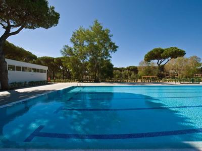 Grande piscine extérieure devant un bâtiment d'hôtel moderne sous un ciel bleu avec des arbres autour.