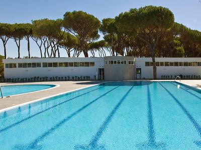 Une grande piscine avec de l'eau claire devant un bâtiment blanc et de grands arbres en arrière-plan.