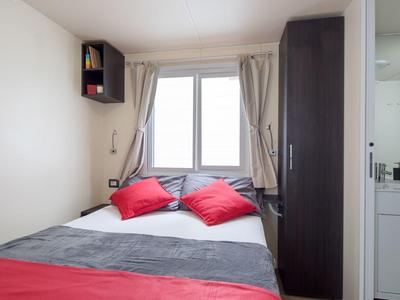 Kleine slaapkamer met tweepersoonsbed, rood beddengoed en kussens, kledingkast en raam met gordijnen.