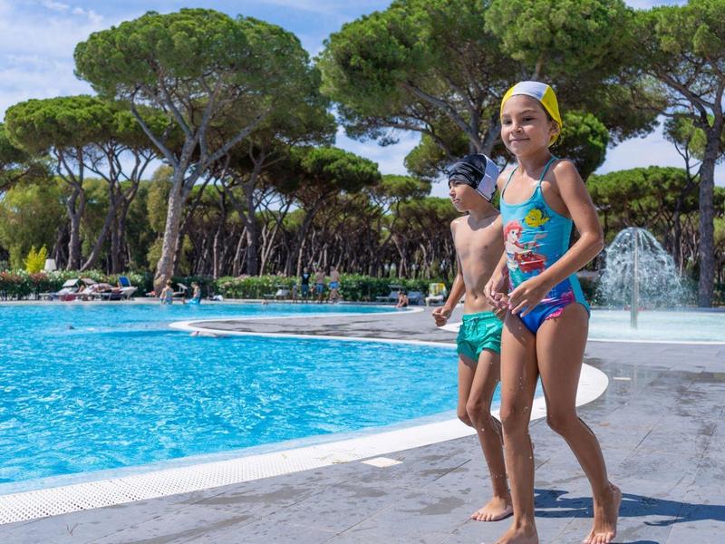 Twee kinderen in zwemkleding lopen langs het zwembad met bomen op de achtergrond.