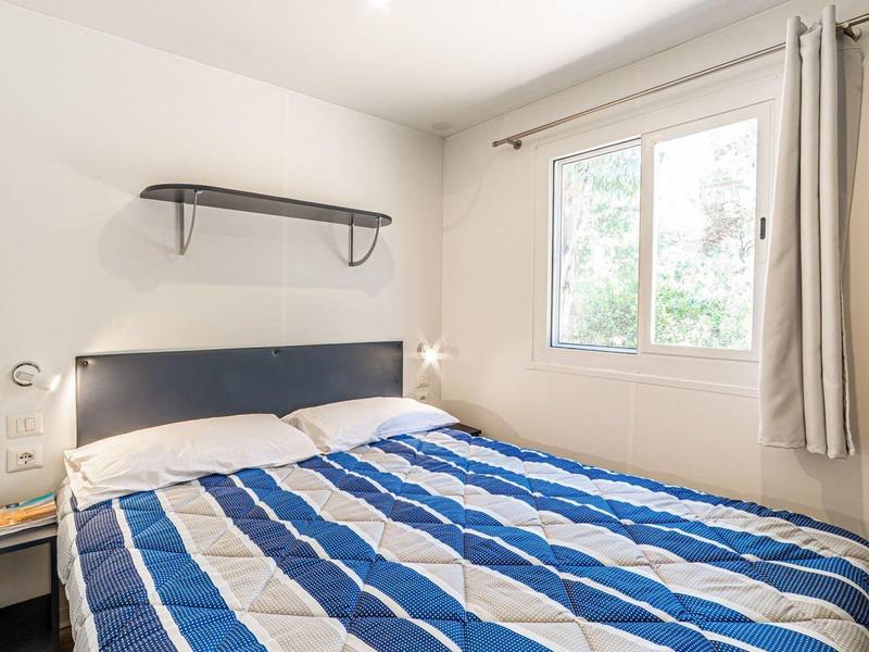 Lichte slaapkamer met tweepersoonsbed en blauw-wit gestreept dekbed naast raam met gordijnen.