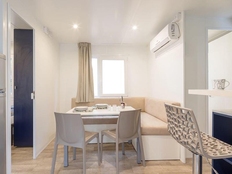 Lichte eetruimte met ronde houten tafel, stoelen, koelkast, airconditioning en groot raam.