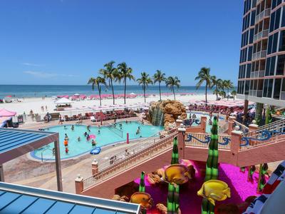 Hotelpoolbereich mit Palmen, Liegestühlen, Gästen und Blick auf den Strand und das Meer unter blauem Himmel.