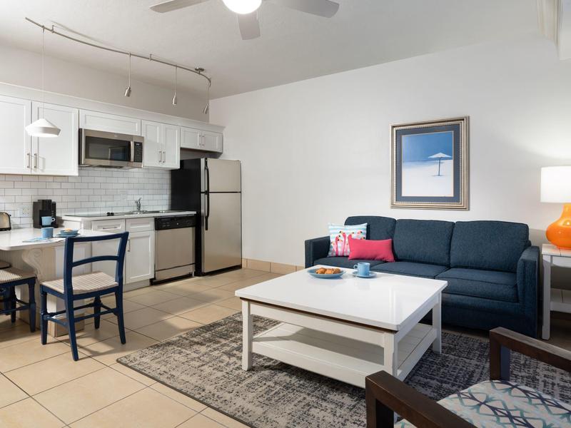 Helle Wohnküche mit moderner weißer Küche, blauer Couch, springender Lampe und Teppich auf Fliesen.