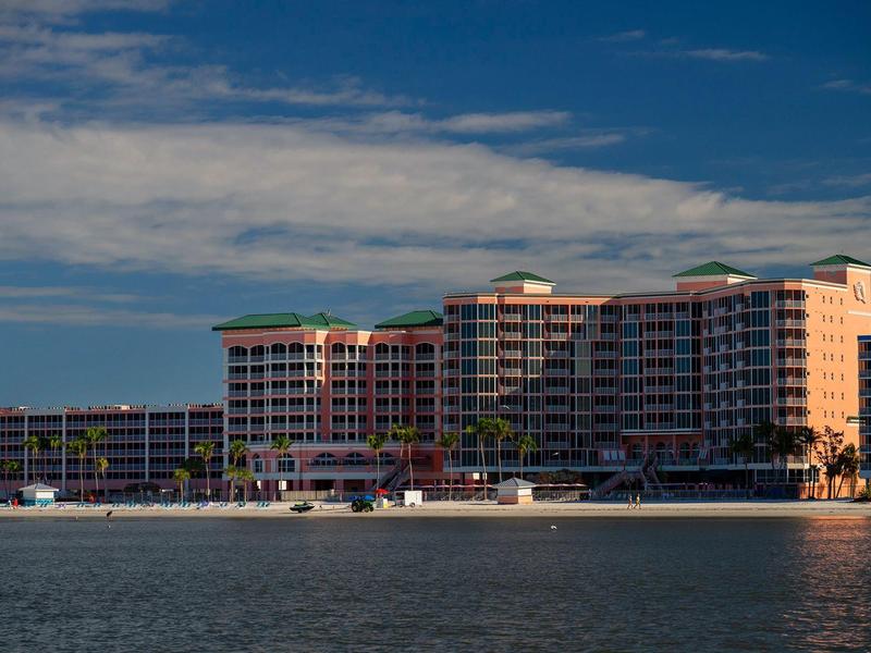 Mehrstöckiges Hotel am Ufer mit blauem Himmel und ruhigem Wasser im Vordergrund.