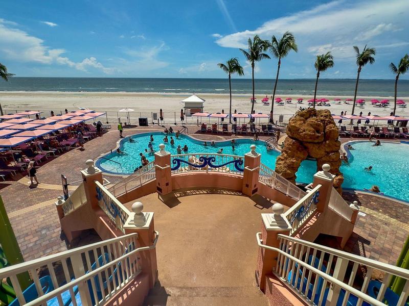 Strand mit Palmen, Poollandschaft, rosa Sonnenschirmen und blauem Himmel.