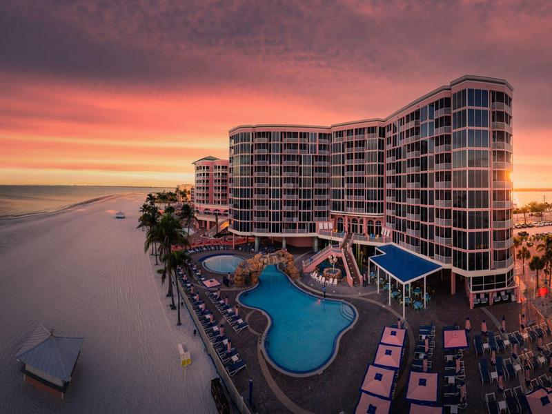 Großes Hotel am Strand mit Pool, Liegestühlen und farbigem Himmel bei Sonnenuntergang.