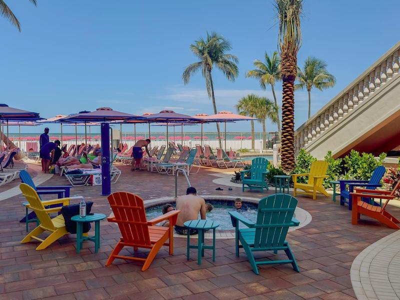 Bunte Stühle und Tische auf Terrasse mit Palmen, Sonnenschirmen und blauen Himmel am Strand.