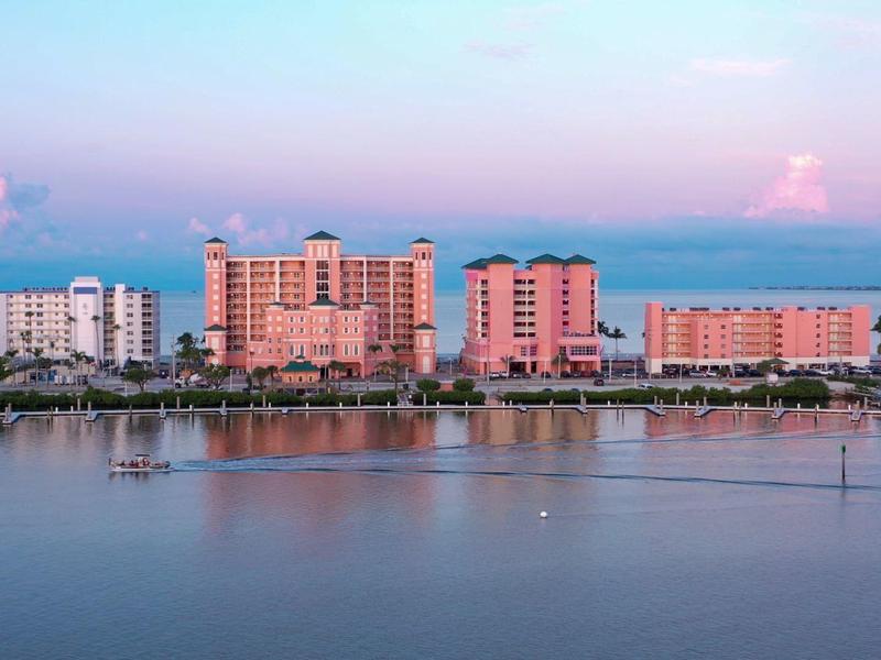 Mehrere orange-rosa Gebäudekomplexe am Wasser mit ruhigem Meer und blauem Himmel im Hintergrund.
