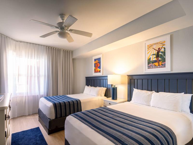 Hell eingerichtetes Schlafzimmer mit zwei Doppelbetten, blauen Kopfstützen und gestreiften Bettdecken.