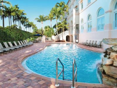 Außenpool mit blauer Wasserfläche, Liegestühlen und Palmen vor einem hellen Gebäude.