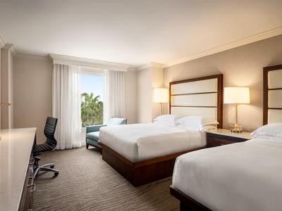 Modern eingerichtetes Hotelzimmer mit zwei Doppelbetten, Schreibtisch und großem Fenster mit Vorhängen.