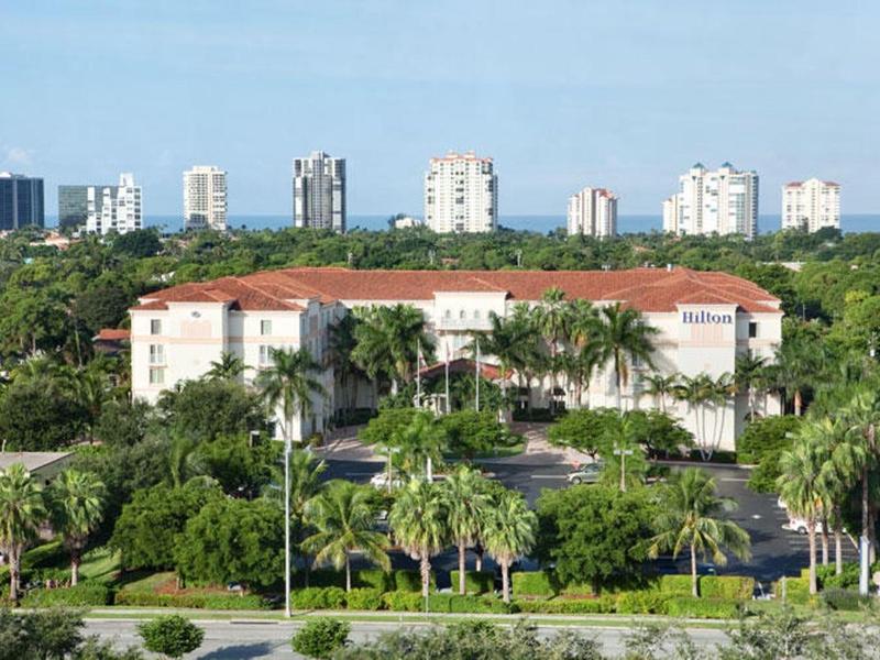 Gebäude mit rotbraunem Dach, umgeben von Palmen und Grünflächen, im Hintergrund Wolkenkratzer.