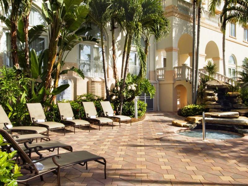 Sonnige Terrasse mit Liegestühlen, Palmen und Springbrunnen vor weißem Gebäude mit Säulen.