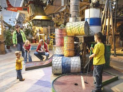Kinder spielen Minigolf in einer bunt dekorierten Indoor-Umgebung mit Fässern und Industriemotiven.