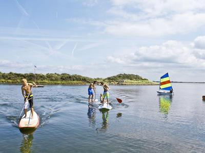 Drei Personen paddeln auf Stand-up-Paddle-Boards auf ruhigem Wasser mit segelndem Boot im Hintergrund.