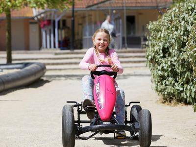 Mädchen fährt mit einem roten Tretfahrzeug auf einer gepflasterten Fläche bei Sonnenschein.
