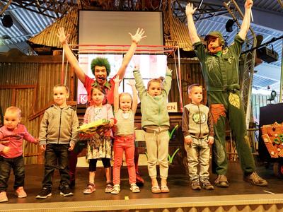 Sechs Kinder und zwei Erwachsene stehen auf einer Bühne und heben die Arme, umgeben von einer bunten Kulisse.