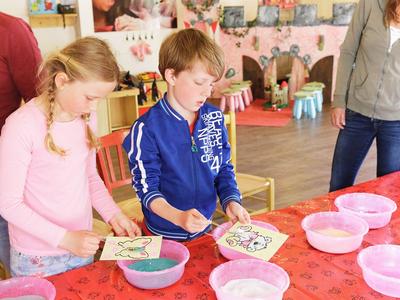 Zwei Kinder bemalen Teller an einem roten Tisch mit Schalen und Pinseln.