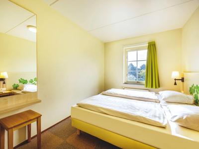 Ein schlicht eingerichtetes Hotelzimmer mit Doppelbett, Fenster mit grünem Vorhang und Holzmöbeln.