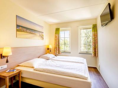 Helles Hotelzimmer mit Doppelbett, Nachttisch, Wandlampen und Fenster mit Vorhängen.