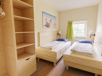 Helles Hotelzimmer mit zwei Einzelbetten, hellem Holz und Fenster mit grüner Gardine.