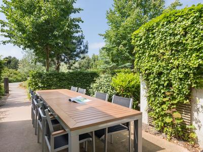 Ein Holztisch mit Stühlen steht draußen auf einer Terrasse, umgeben von grünen Hecken und Pflanzen.