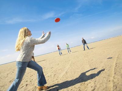 Person wirft Frisbee an einem sonnigen Strand mit mehreren Menschen im Hintergrund.