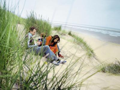 Zwei Personen sitzen in Dünen am Strand und genießen die Aussicht bei bewölktem Himmel.