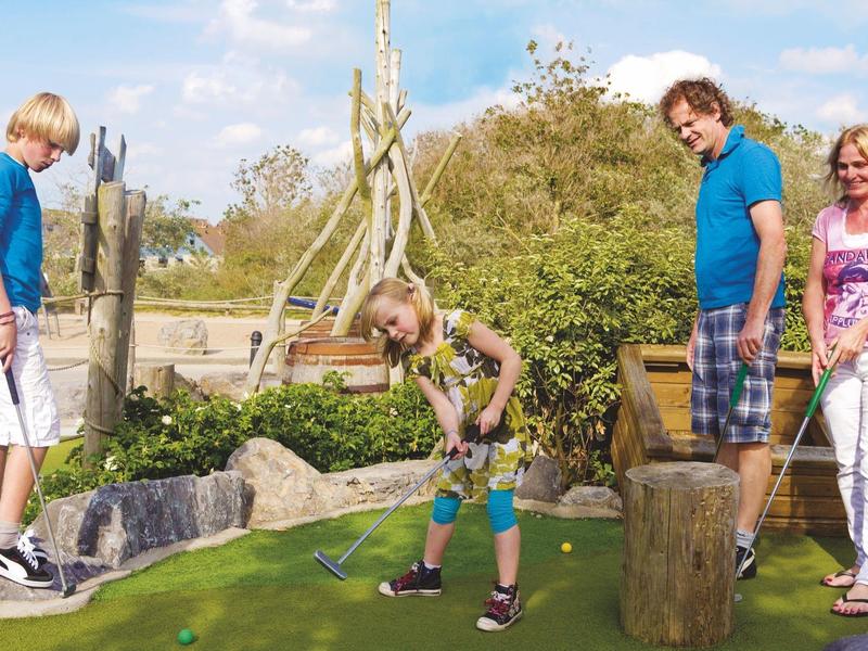 Vier Personen spielen Minigolf im Freien auf grünem Kunstrasen mit Holz- und Felsdekoration.