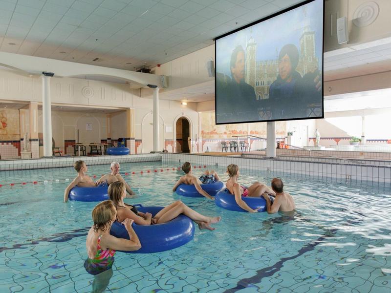 Kinder im Hallenbad sehen animierten Film auf großer Leinwand über dem Becken.