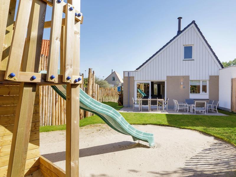 Kinderspielplatz mit Rutsche aus Holz und modernem weißem Haus im Hintergrund bei klarem Himmel.