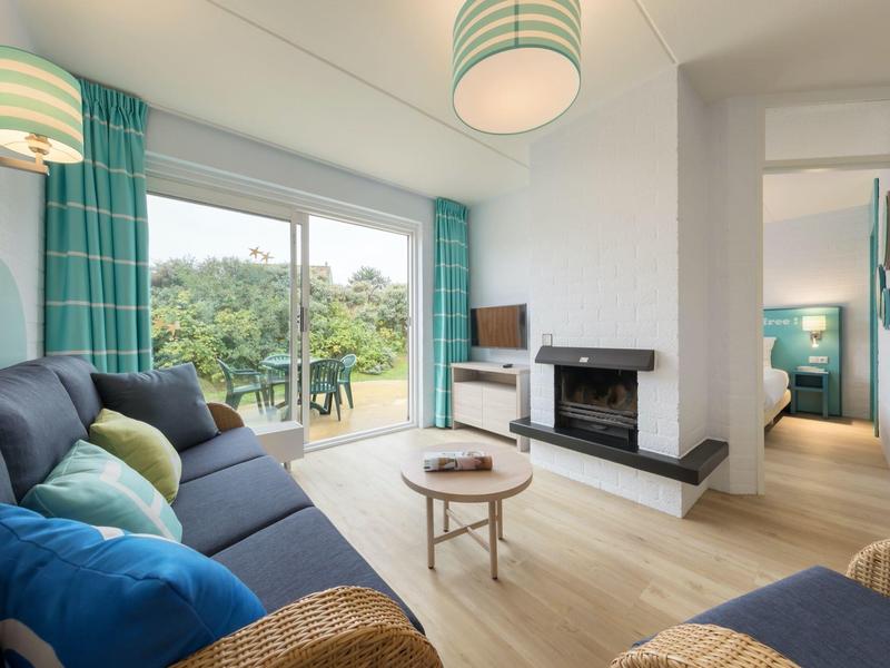 Heller Wohnzimmerbereich mit großem Fenster, Kamin, blauen Sofas und Holzfußboden, offen zum Flur.