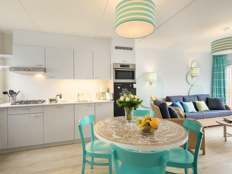 Helles offenes Wohn- und Esszimmer mit türkisen Stühlen, Rundtisch und modernem Küchenbereich.