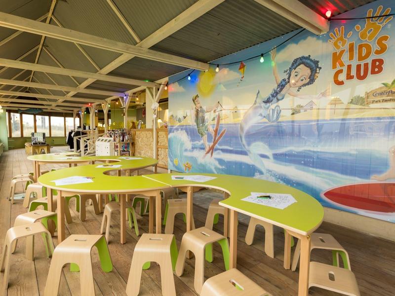 Heller Kinderraum mit buntem Wandbild, geschwungenen Tischen und kleinen Hockern aus Kunststoff.