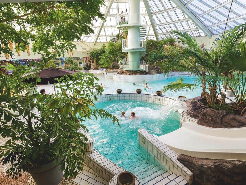 Innenliegendes Schwimmbad mit runden Pools, Pflanzen und robustem Glasdach, viele Besucher schwimmen.