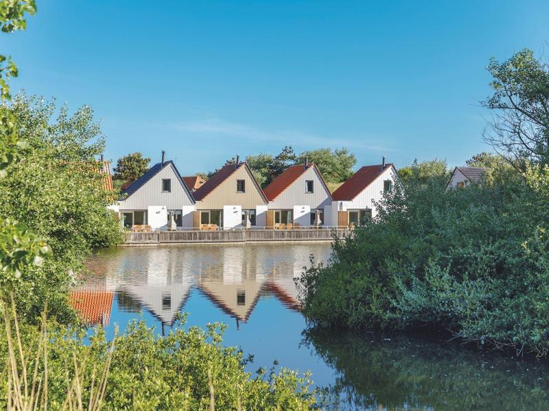 Vier weiße Häuser mit roten Dächern spiegeln sich im ruhigen Fluss, umgeben von grünen Büschen.