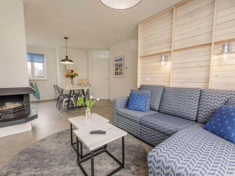 Gemütliches Wohnzimmer mit grauer Eckcouch, Kamin, weißem Tisch und Essbereich im Hintergrund.