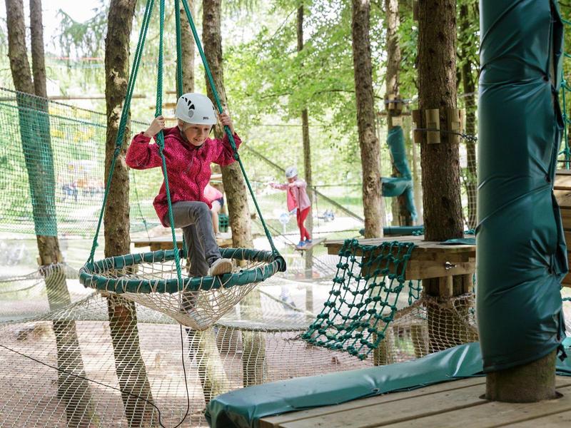 Niño trepando con casco y arnés de seguridad en parque de cuerdas altas entre árboles en bosque hotelero.