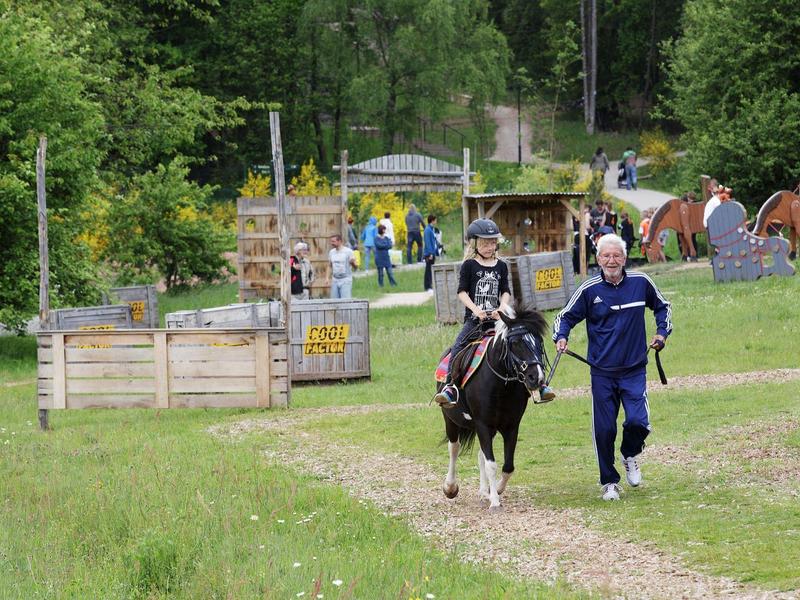 Un niño monta un pony acompañado por un adulto en un camino forestal con barreras de madera al fondo.